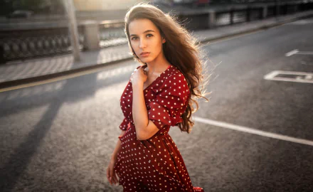 2K Quad HD PC desktop wallpaper: brunette model with long hair in a red polka-dot dress standing on an urban street, backlit by warm evening light, elegant background