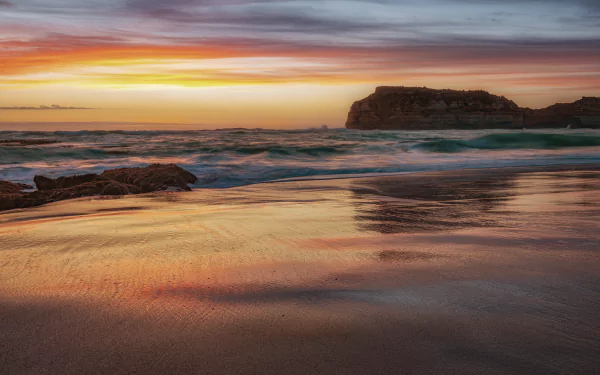 4K Ultra HD desktop wallpaper showcasing a serene beach at sunset with gentle ocean waves and a rocky cliff under a colorful sky.