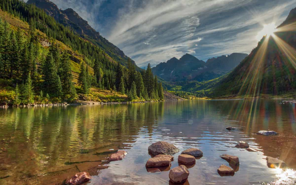Sunbeam breaks through clouds over a tranquil Colorado mountain lake, reflecting stone shores and dense forest under a vibrant sky in this HD nature wallpaper.