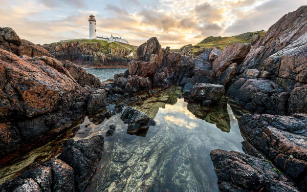 5K Ultra HD PC desktop wallpaper: Irish coastline with a man-made lighthouse on a rocky headland, sunset sky reflected in a tidal pool among jagged rocks.