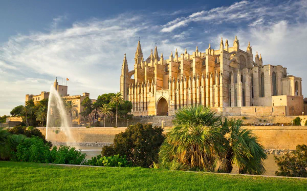 HD PC desktop wallpaper of a religious cathedral in Mallorca, Spain, with a fountain, palm trees and green lawns under a bright blue sky.