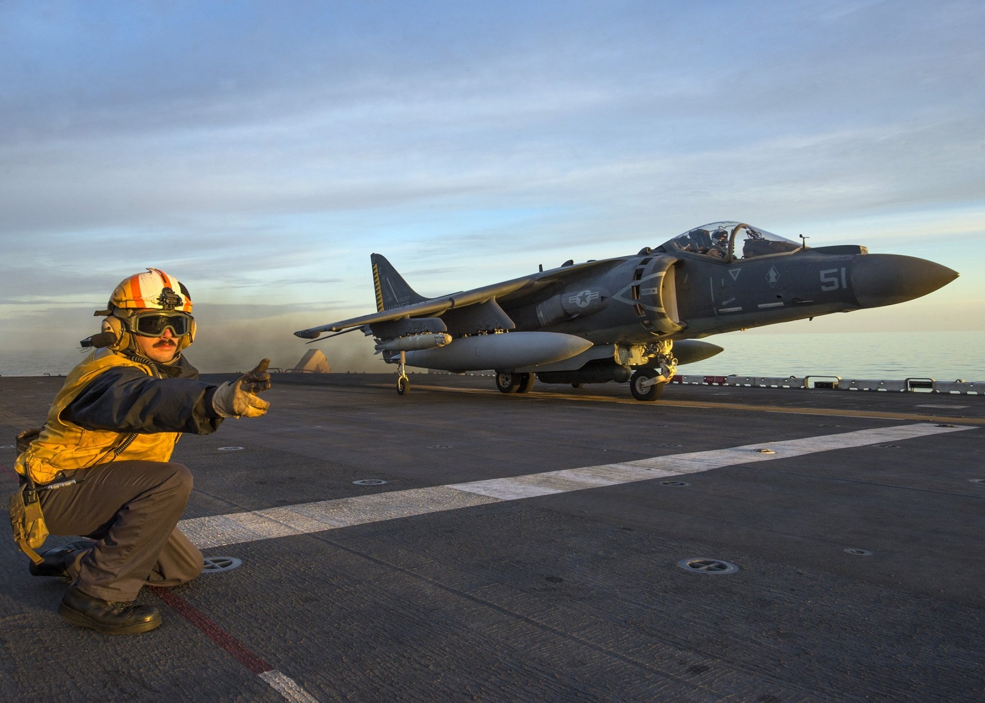 HD desktop wallpaper featuring a McDonnell Douglas AV-8B Harrier II jet fighter warplane on a military aircraft carrier deck during sunset.