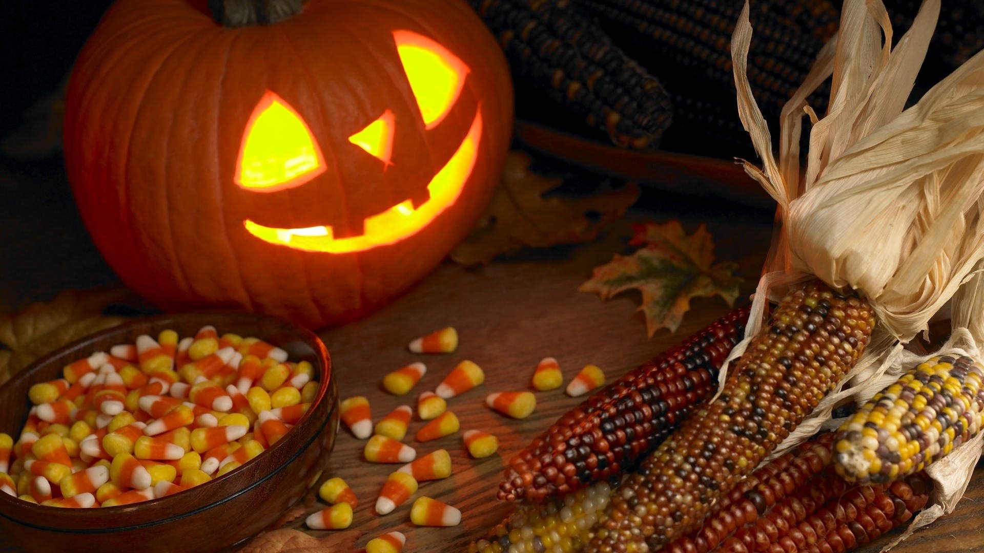 HD desktop wallpaper featuring a glowing carved pumpkin, colorful candy corn, and multicolored ornamental corn evokes a festive Halloween holiday atmosphere.