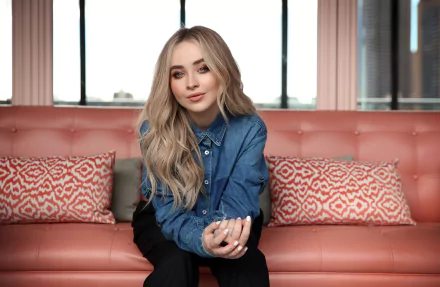 American actress and singer Sabrina Carpenter with long blonde hair and blue eyes, seated on a pink sofa in a modern indoor setting.