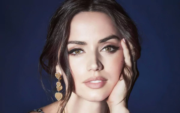 Close-up 4K portrait of Cuban actress Ana de Armas with green eyes, brunette hair, and heart-shaped earrings against a dark blue background.