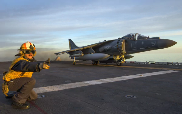 HD desktop wallpaper featuring a McDonnell Douglas AV-8B Harrier II jet fighter warplane on a military aircraft carrier deck during sunset.