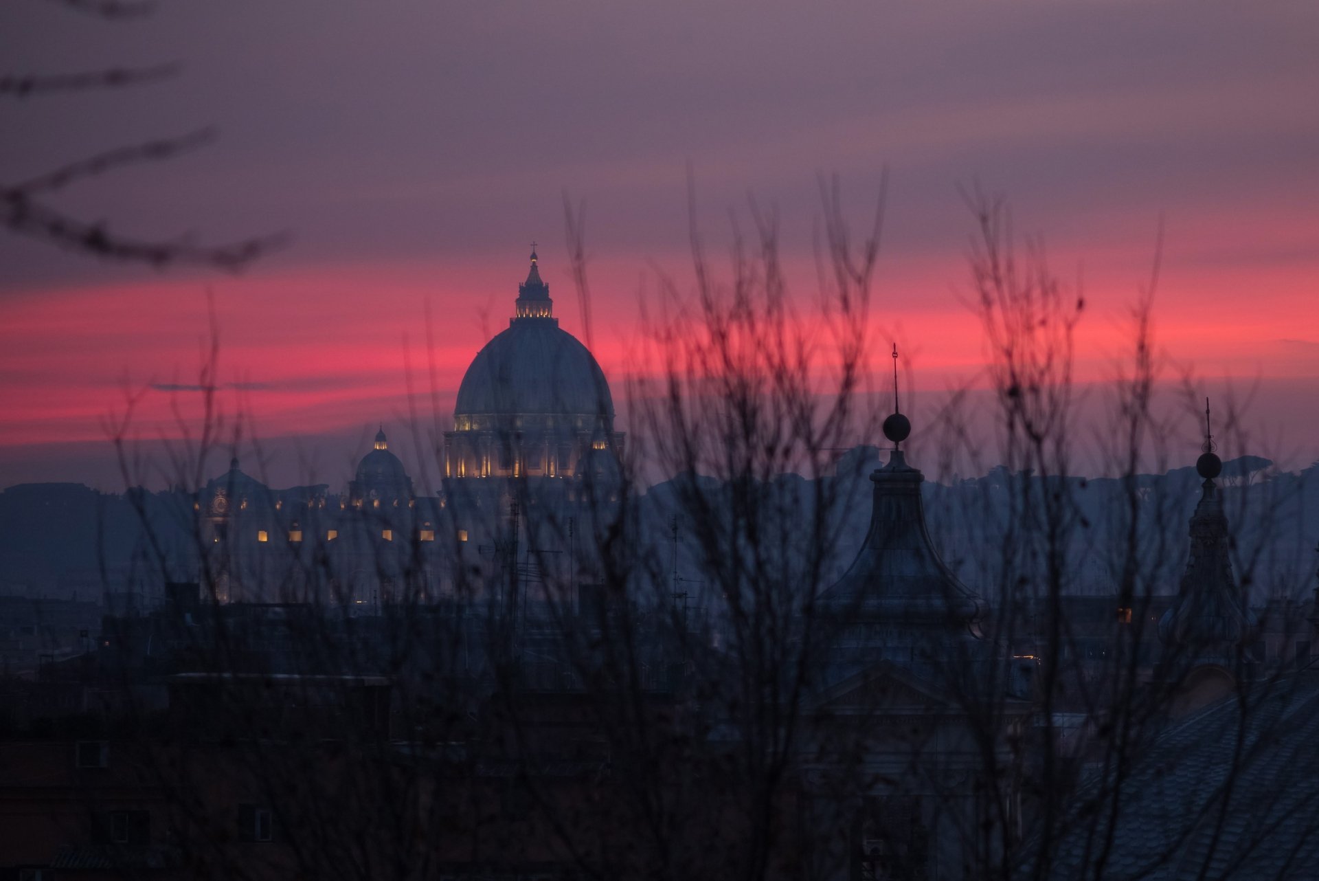 HD desktop wallpaper: St. Peter's Basilica silhouetted against a pink-purple evening sky over the Vatican in Rome, with bare branches in the foreground.