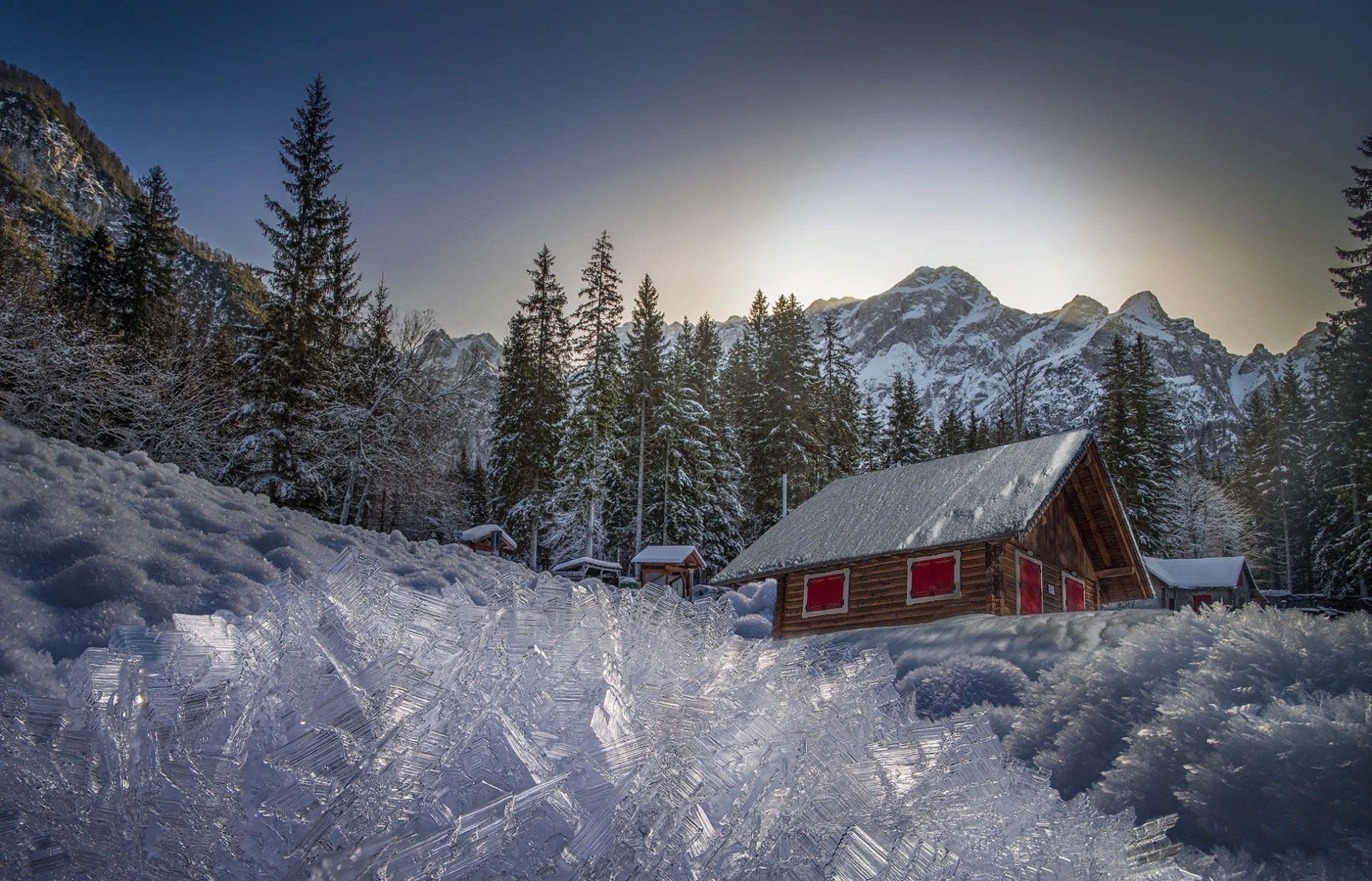 HD PC desktop wallpaper photograph: snow-covered cabin and fir trees beneath snowy mountain peaks, red house in serene winter nature.