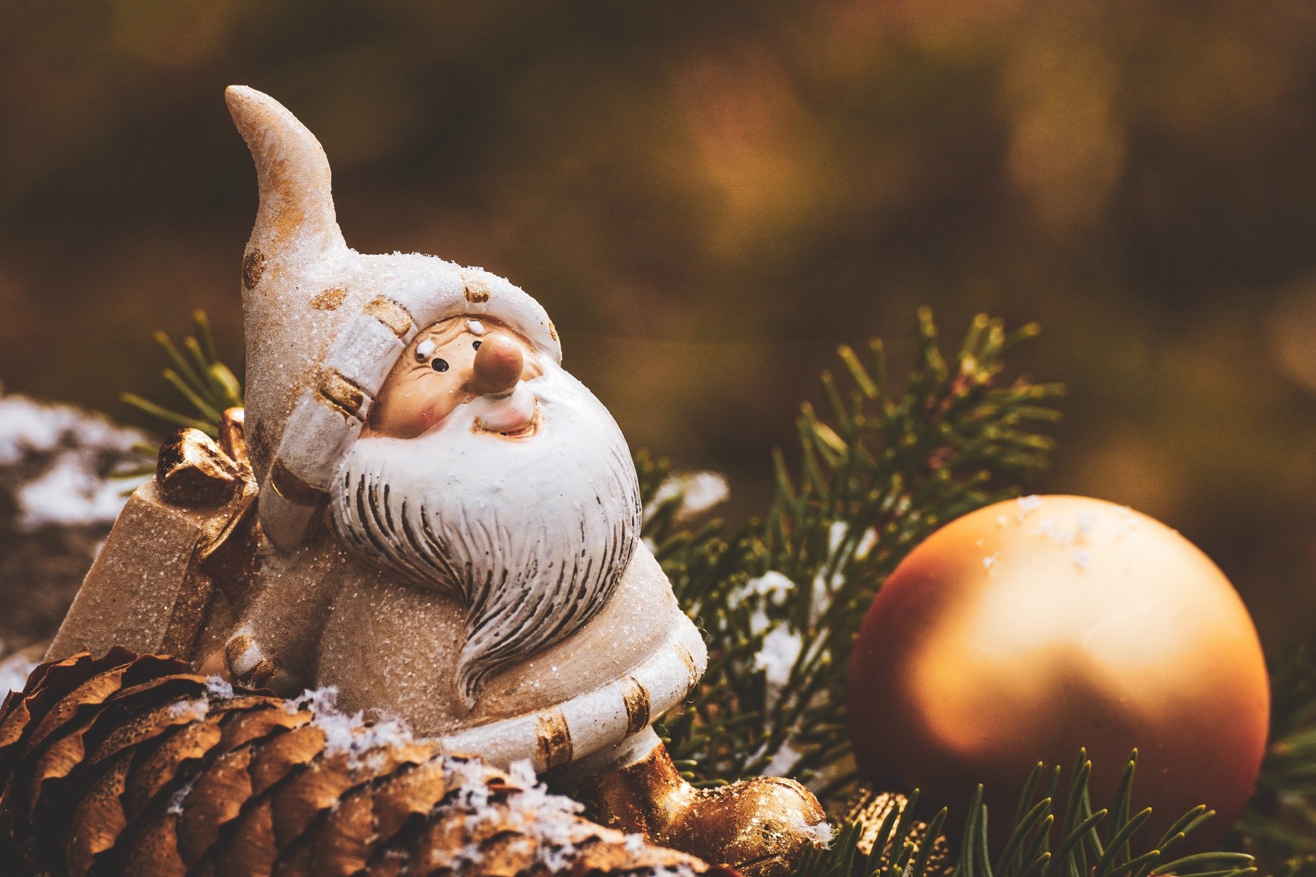 Holiday Christmas 4K Ultra HD PC desktop wallpaper: cozy Santa figurine among snowy pinecones and evergreen branches with a gold ornament and warm bokeh background.