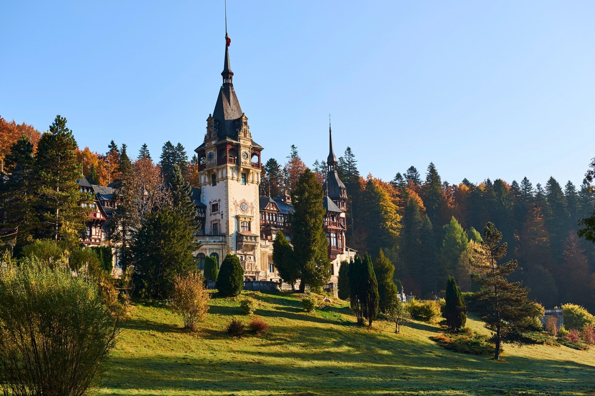 Peles Castle in Transylvania, Romania, stands majestically surrounded by autumn trees under a clear blue sky, featured in an HD PC desktop wallpaper.