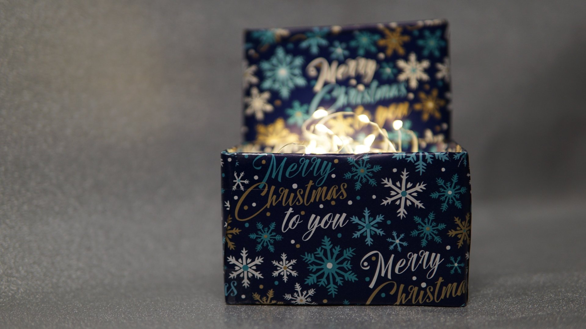 5K Ultra HD PC desktop wallpaper: close-up of two Christmas gift boxes wrapped in navy snowflake paper reading Merry Christmas, with soft string lights creating warm bokeh holiday glow.