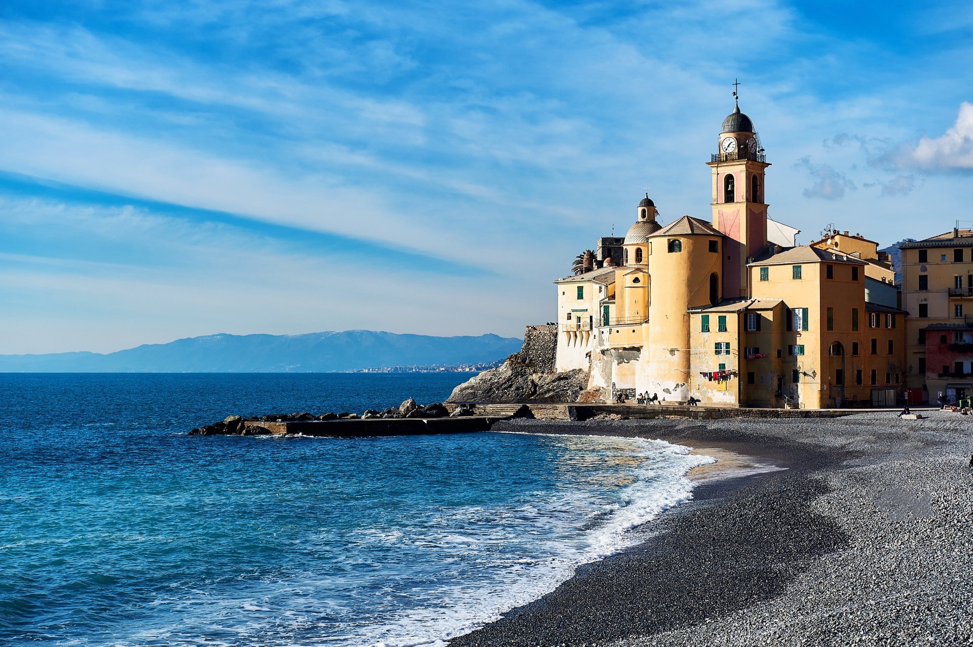 4K Ultra HD desktop wallpaper of Camogli, Liguria: pastel church and coastal houses on a rocky beach, waves lapping the pebbled shore under a bright blue sky.