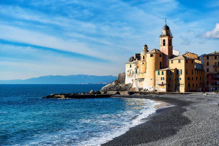 4K Ultra HD desktop wallpaper of Camogli, Liguria: pastel church and coastal houses on a rocky beach, waves lapping the pebbled shore under a bright blue sky.