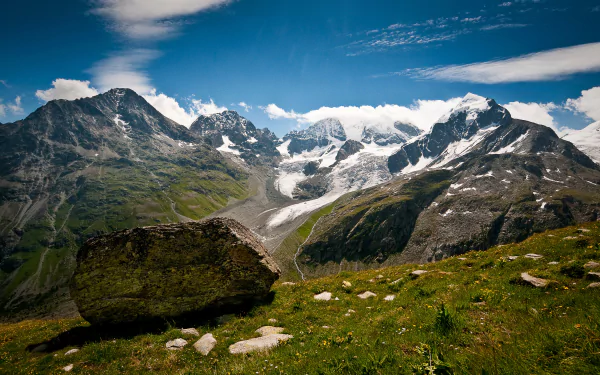 4K Ultra HD PC desktop wallpaper: Swiss Alps panorama of alpine nature with jagged mountain peaks, glacier-fed valley, green foreground and boulder under a bright blue sky.