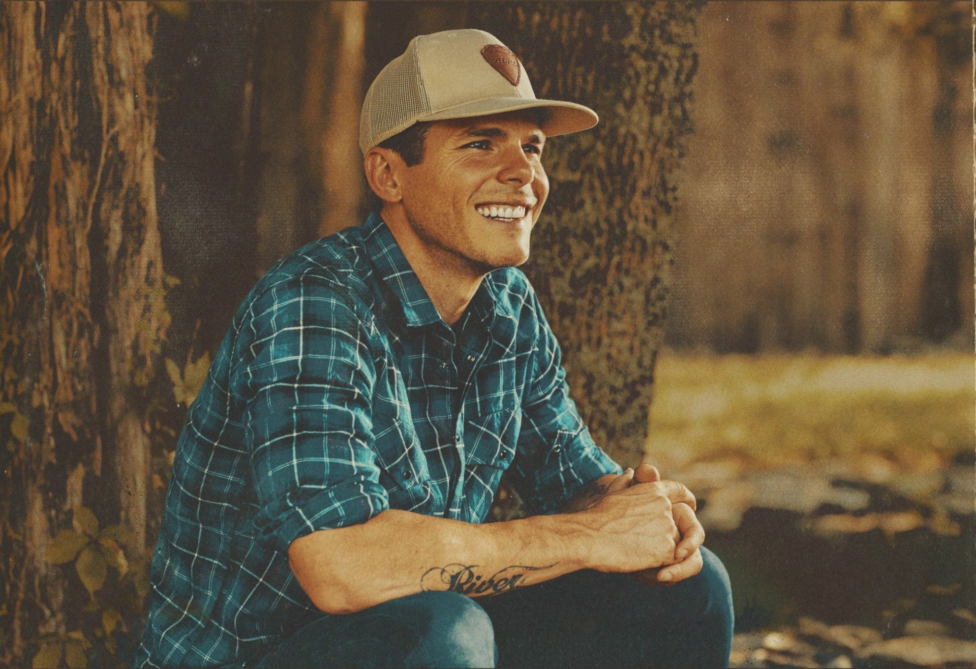 HD desktop wallpaper featuring Granger Smith in a blue plaid shirt and hat, smiling while seated outdoors against a tree.