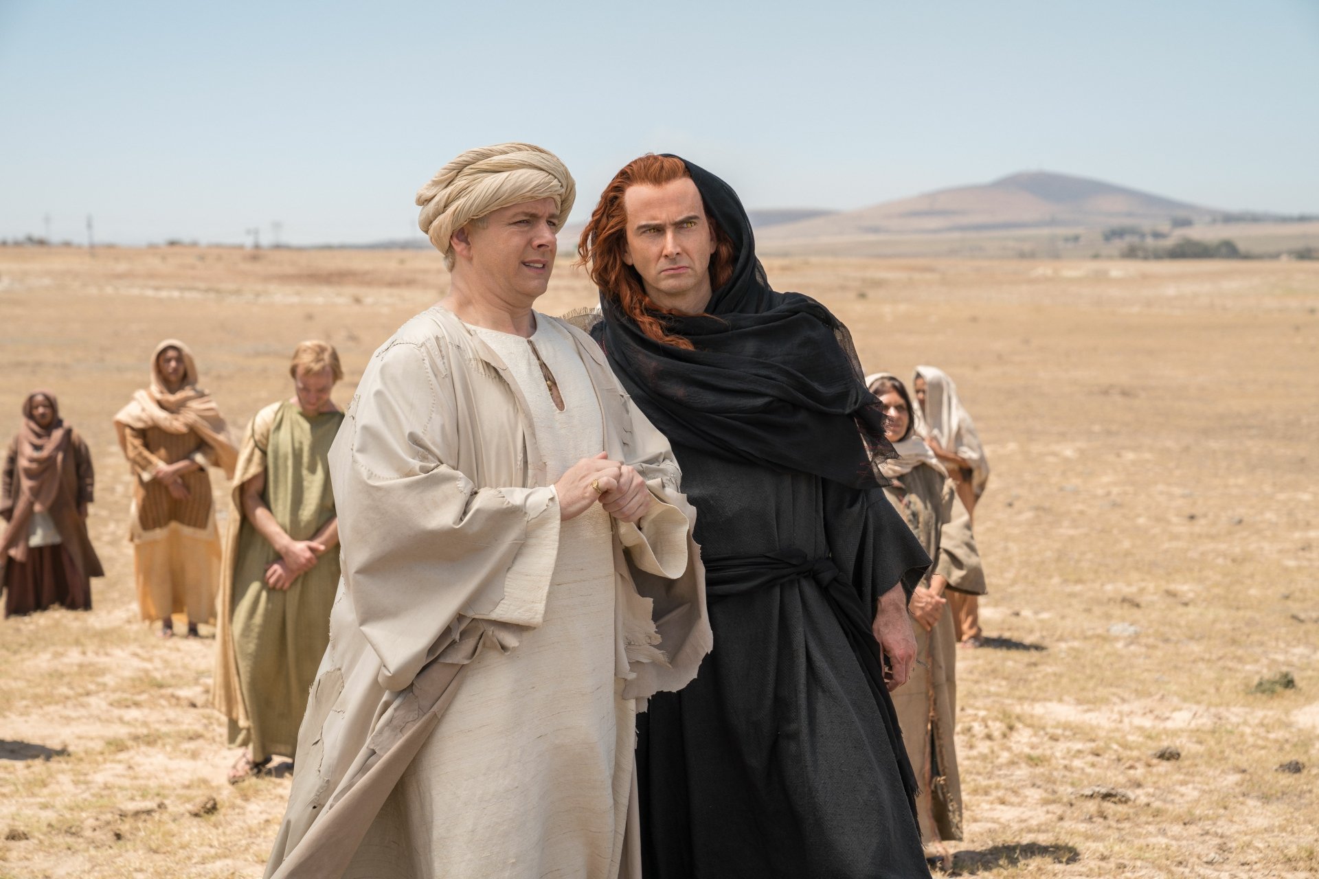 David Tennant as Crowley and Michael from Good Omens stand in a desert landscape, dressed in period attire, with others in the background in this 8K Ultra HD TV show wallpaper.