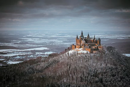 Hohenzollern Castle perched on a snowy mountain ridge under a cloudy winter sky, overlooking a vast landscape and distant horizon.