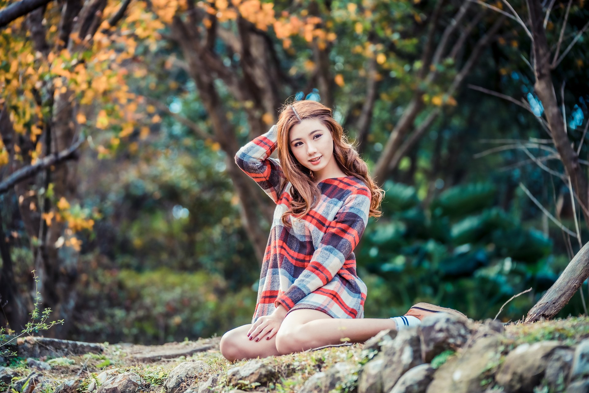 A brunette Asian woman model sits on a rock in nature, surrounded by blurred autumn foliage, captured with depth of field in 4K Ultra HD quality.