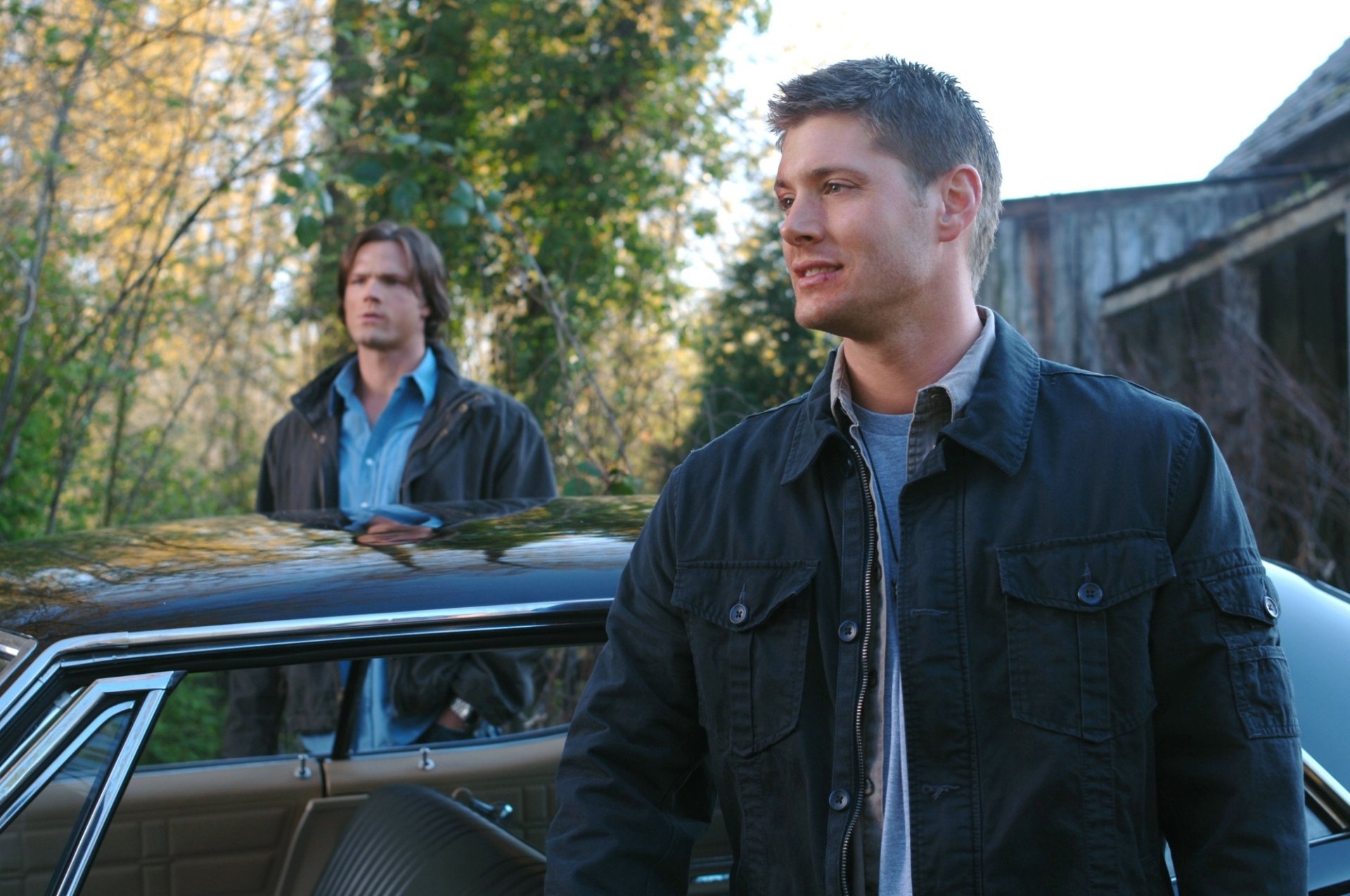 HD PC desktop wallpaper: two men in jackets beside a classic car in a wooded driveway, a background image from the TV show Supernatural.