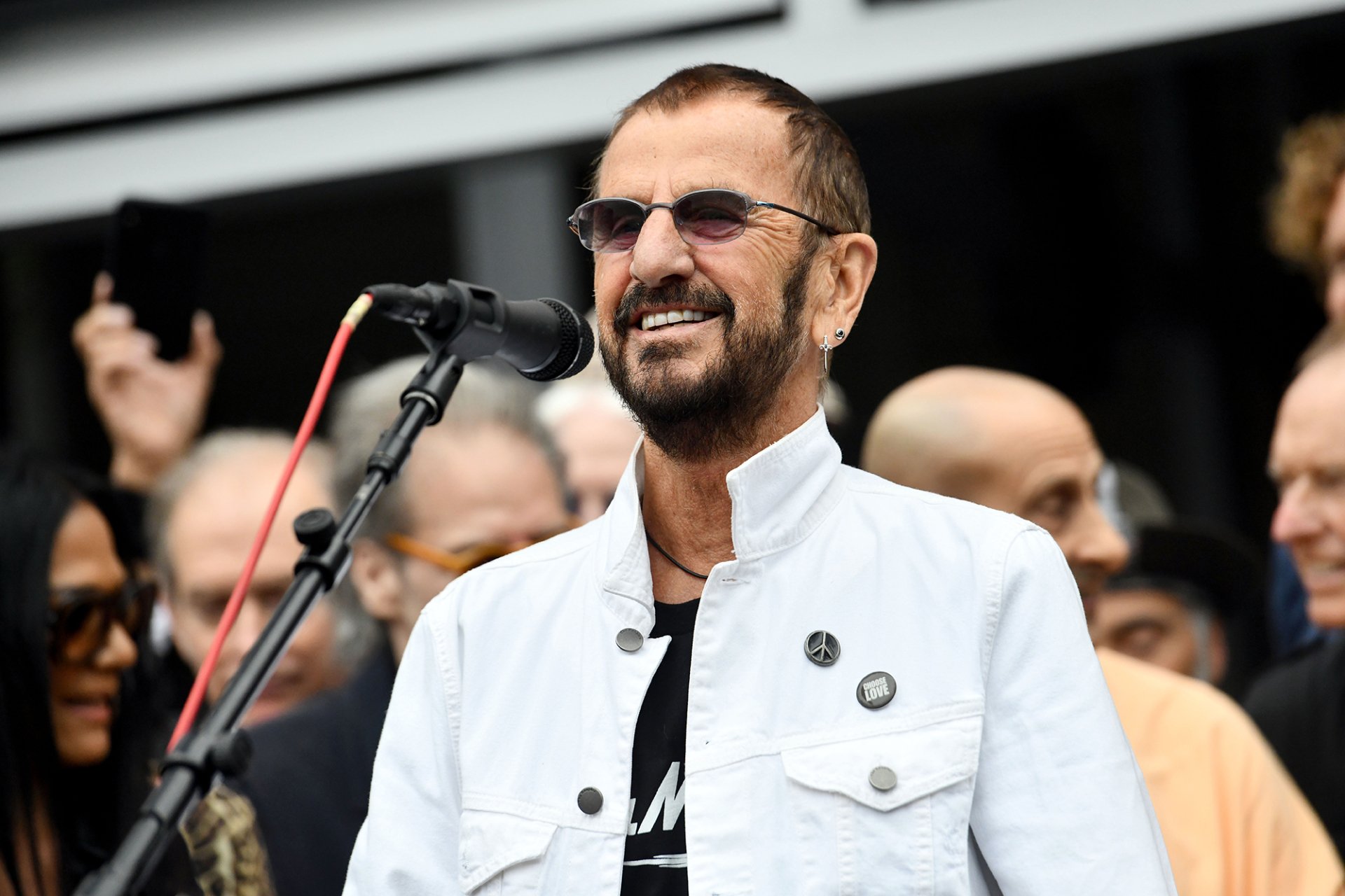 HD desktop wallpaper featuring a person standing at a microphone with a smiling expression in a public setting.
