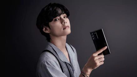 V (Kim Taehyung) of BTS poses against a dark background, holding a sleek phone, creating a striking 4K Ultra HD music-themed PC desktop wallpaper.