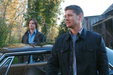 HD PC desktop wallpaper: two men in jackets beside a classic car in a wooded driveway, a background image from the TV show Supernatural.