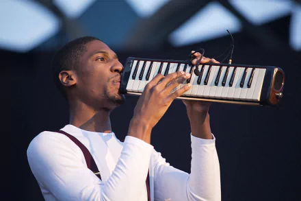 Jon Batiste performs on stage, playing a melodica, in a high-definition desktop wallpaper.