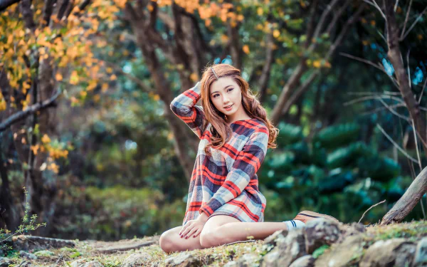 A brunette Asian woman model sits on a rock in nature, surrounded by blurred autumn foliage, captured with depth of field in 4K Ultra HD quality.