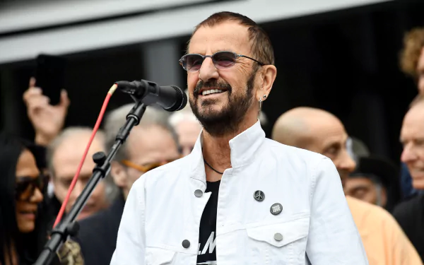 HD desktop wallpaper featuring a person standing at a microphone with a smiling expression in a public setting.