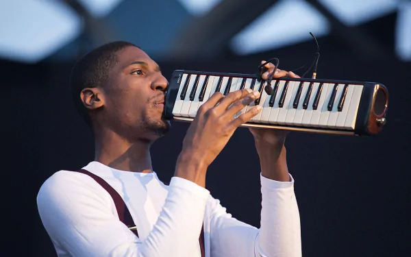 Jon Batiste performs on stage, playing a melodica, in a high-definition desktop wallpaper.