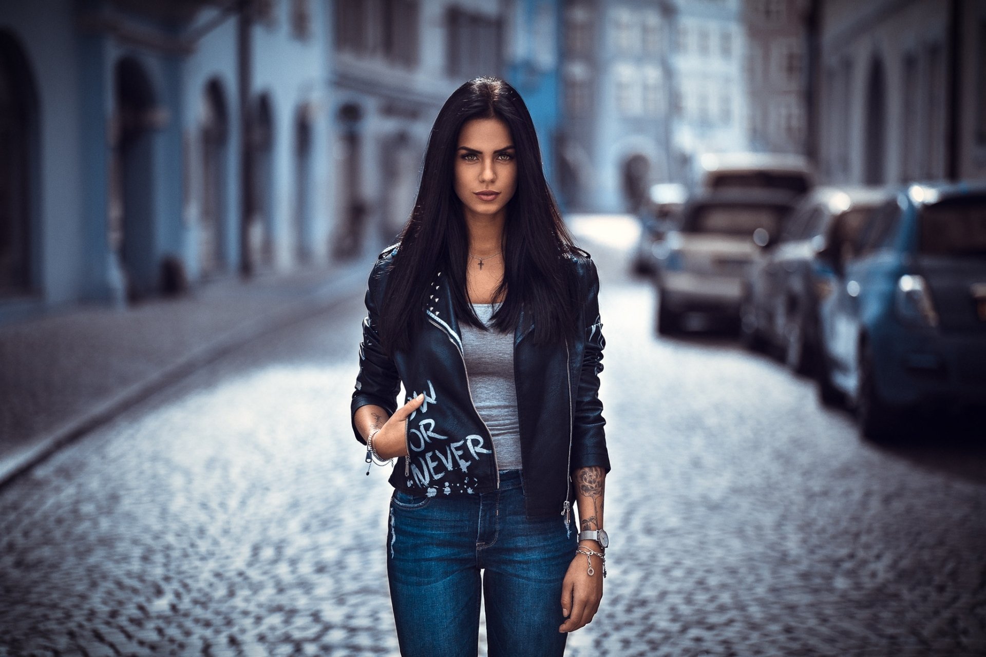 HD desktop wallpaper featuring model Marlen Valderrama Alvaréz with black hair, wearing a leather jacket, standing on a cobblestone street with a blurred background.