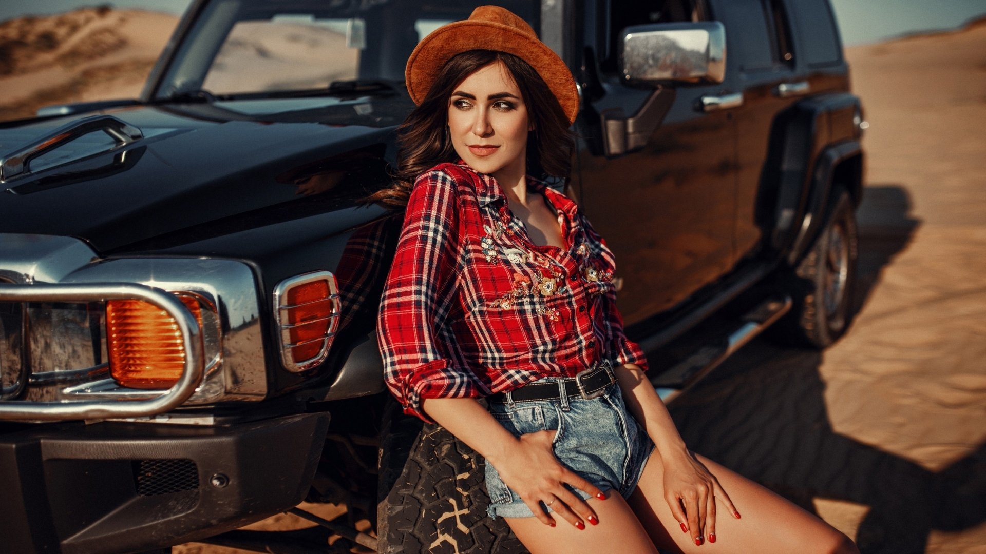 Brunette woman model in a straw hat and plaid shirt with denim shorts leaning against a black SUV in a desert — 2K Quad HD PC desktop wallpaper/background.