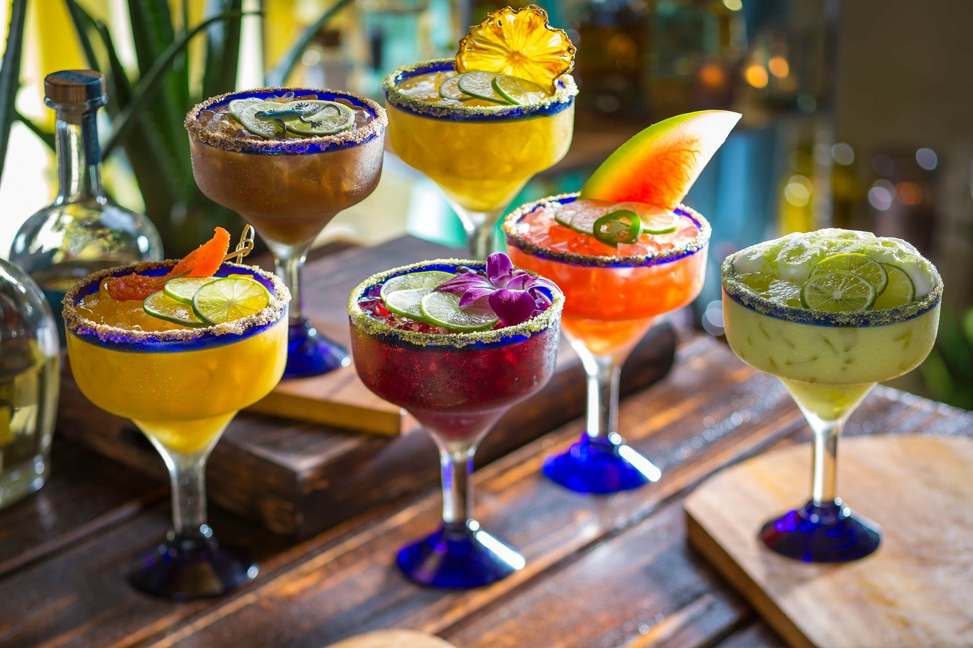 A vibrant array of six colorful margaritas garnished with fruits and herbs, set against a rustic wooden backdrop, creating an enticing food and drink scene. High-definition desktop wallpaper.