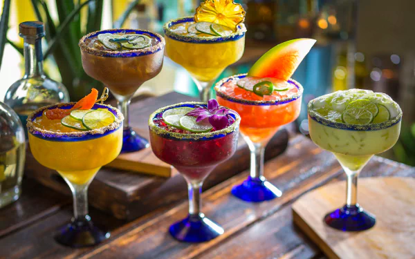 A vibrant array of six colorful margaritas garnished with fruits and herbs, set against a rustic wooden backdrop, creating an enticing food and drink scene. High-definition desktop wallpaper.