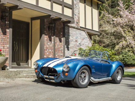 Blue Shelby Cobra 427 (AC Cobra) with white racing stripes parked by a brick Tudor-style house — 4K Ultra HD PC desktop wallpaper.