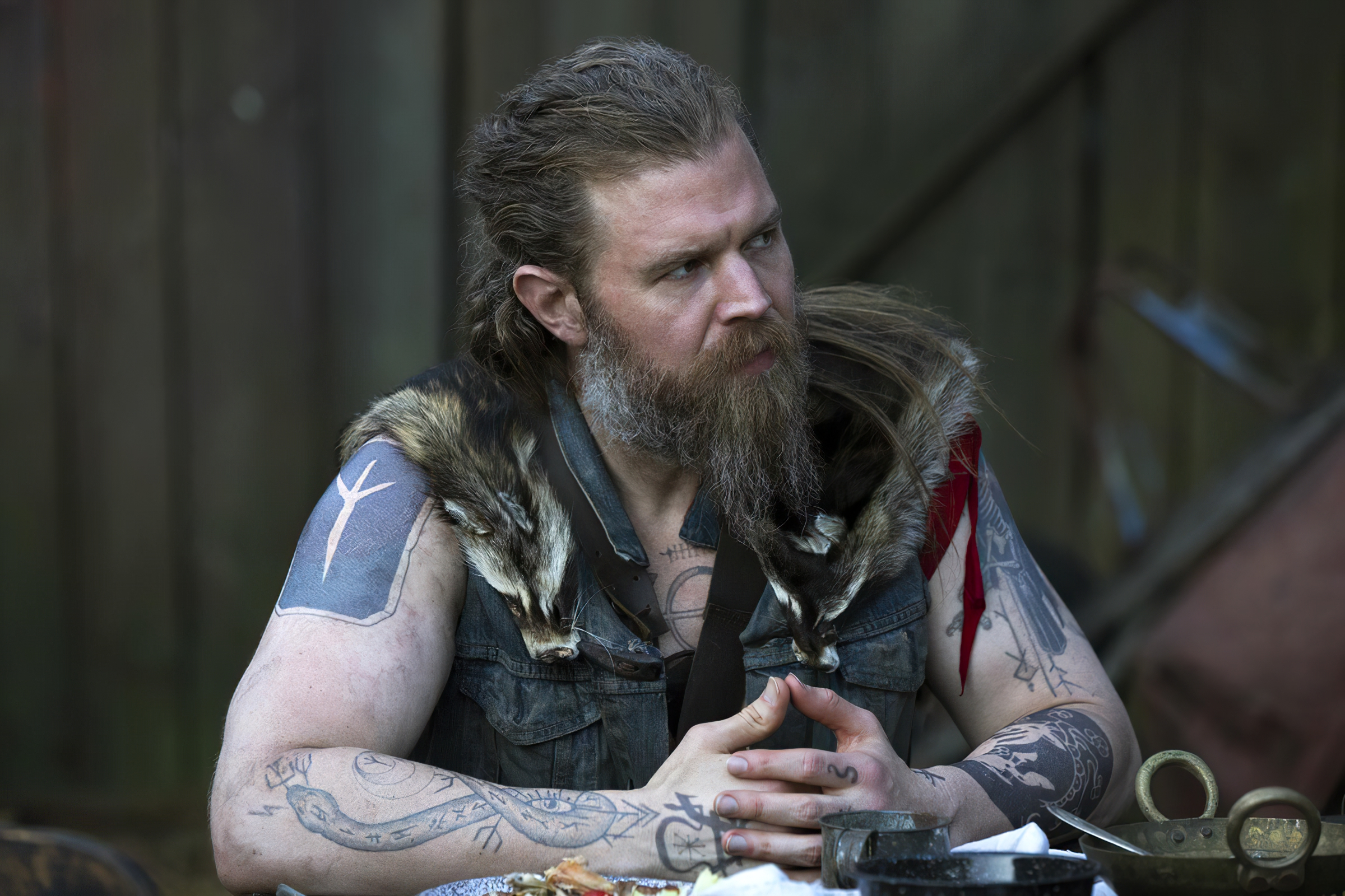HD wallpaper of a character from Outlander, featuring a bearded man with tattoos wearing a vest with fur accents, contemplating thoughtfully.
