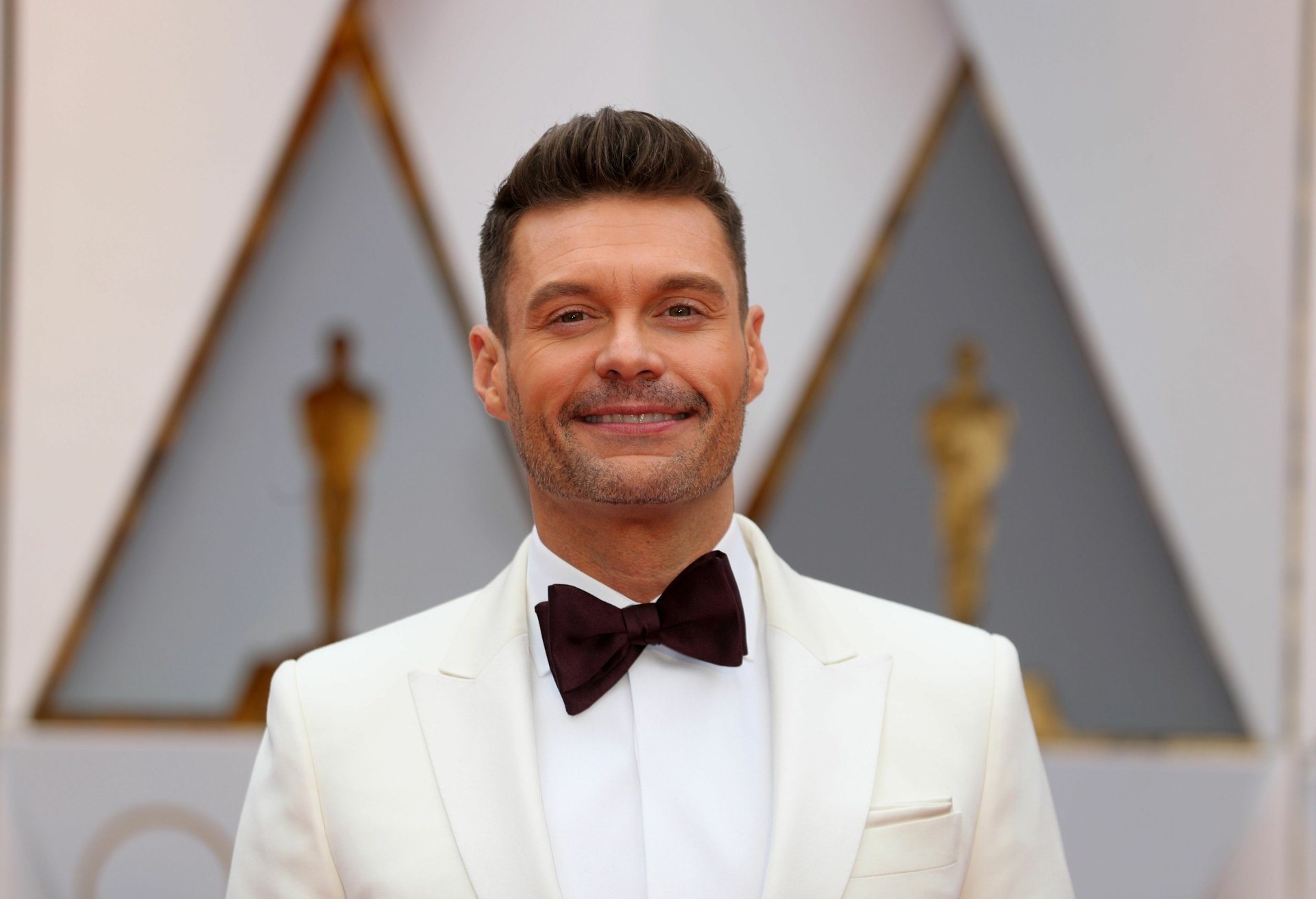 A stylish HD desktop wallpaper featuring Ryan Seacrest, smiling confidently in a white tuxedo, set against a backdrop of iconic Oscar statues.