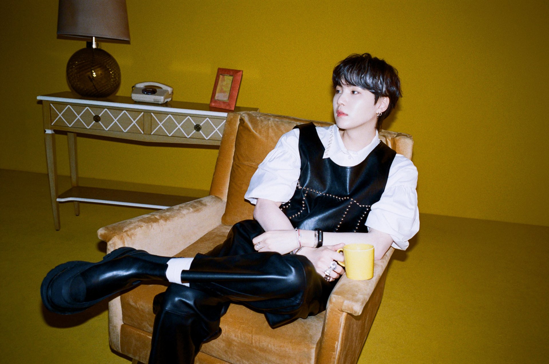 K-pop–styled singer in black vest and white shirt lounges in a mustard armchair, holding a yellow mug; minimalist music-themed 2K Quad HD PC desktop wallpaper and background.