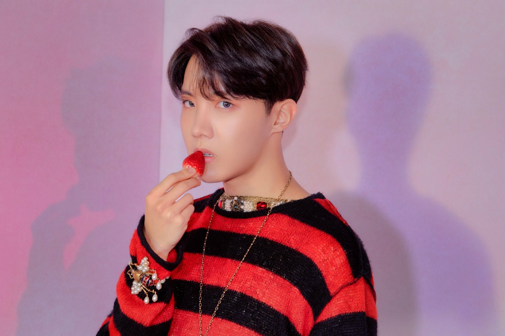 J-Hope of BTS, a K-pop singer, is captured in this vibrant 4K Ultra HD wallpaper, wearing a red and black striped sweater while holding a red object near his lips.