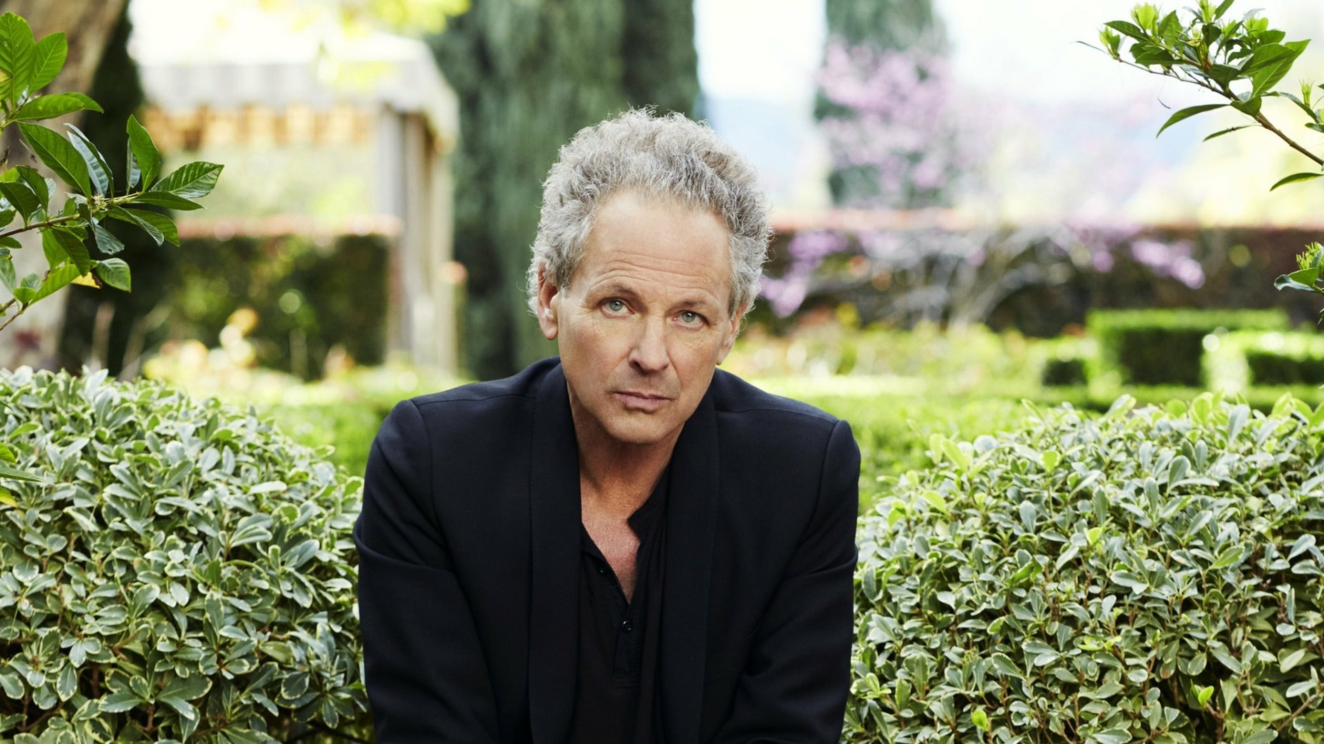 A serene HD desktop wallpaper featuring Lindsey Buckingham, captured in a lush garden setting, exuding a contemplative mood amidst vibrant greenery.
