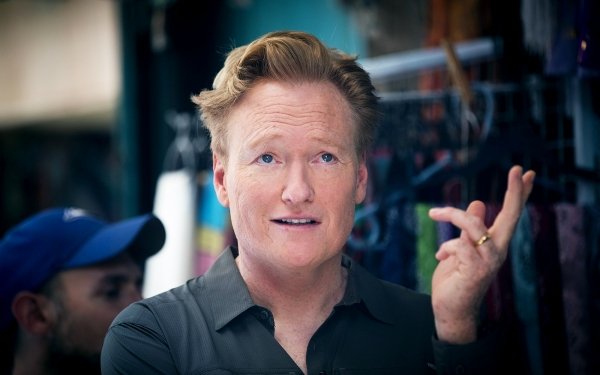 Smiling man with iconic hairstyle gesturing with his hand, in front of a blur market background, ideal for HD desktop wallpaper use.