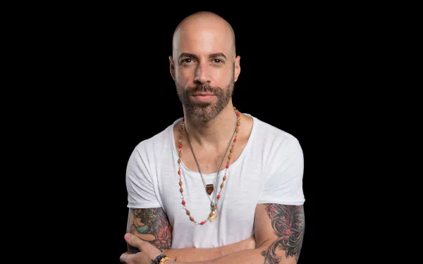 HD wallpaper of a male musician with tattoos, wearing a white t-shirt and a necklace, posing against a black background.