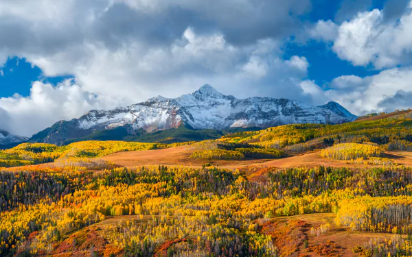 5K Ultra HD PC desktop wallpaper: fall mountain nature landscape with golden aspen meadows, rolling hills and a snow-capped peak under dramatic clouds.
