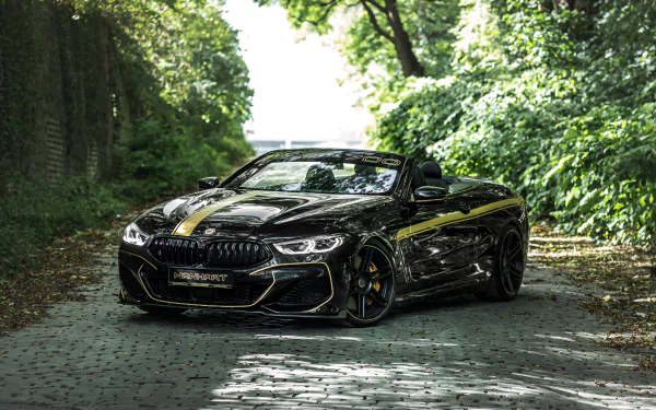 A sleek black Manhart MH8 700 Cabrio, based on the BMW 8 Series, parked on a sunlit cobblestone path surrounded by lush green trees in this 4K Ultra HD desktop wallpaper.