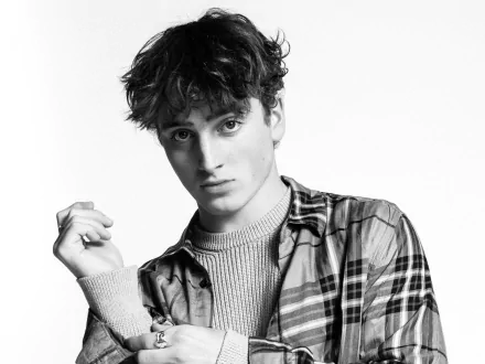 Black-and-white portrait of a young male musician in a plaid shirt and knit sweater, moody 2K Quad HD PC desktop wallpaper with music-themed styling.