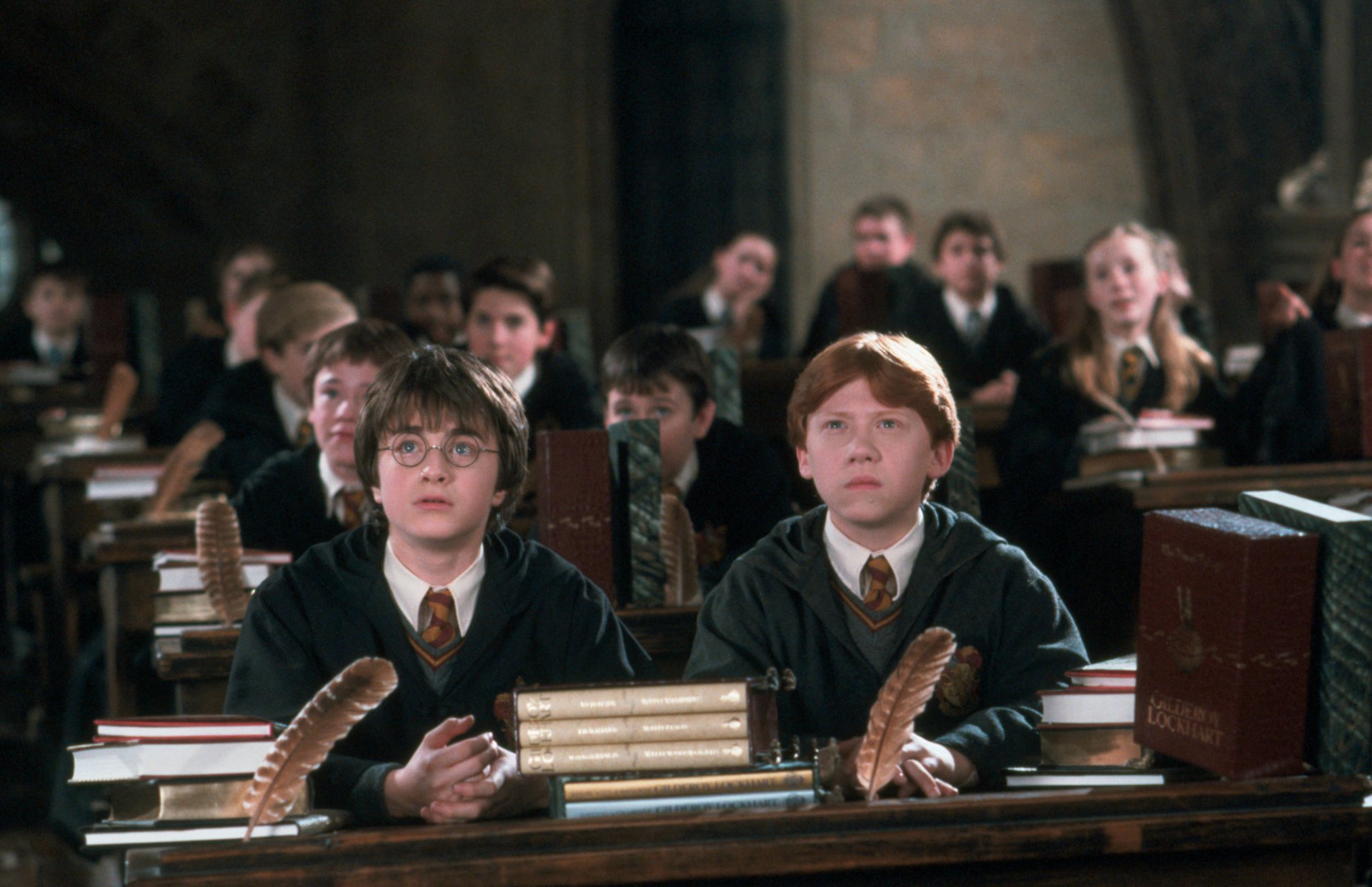 Two young Hogwarts students—one with round glasses, one red-haired—sit at desks with quills and stacked books in a crowded classroom scene from Harry Potter and the Chamber of Secrets.