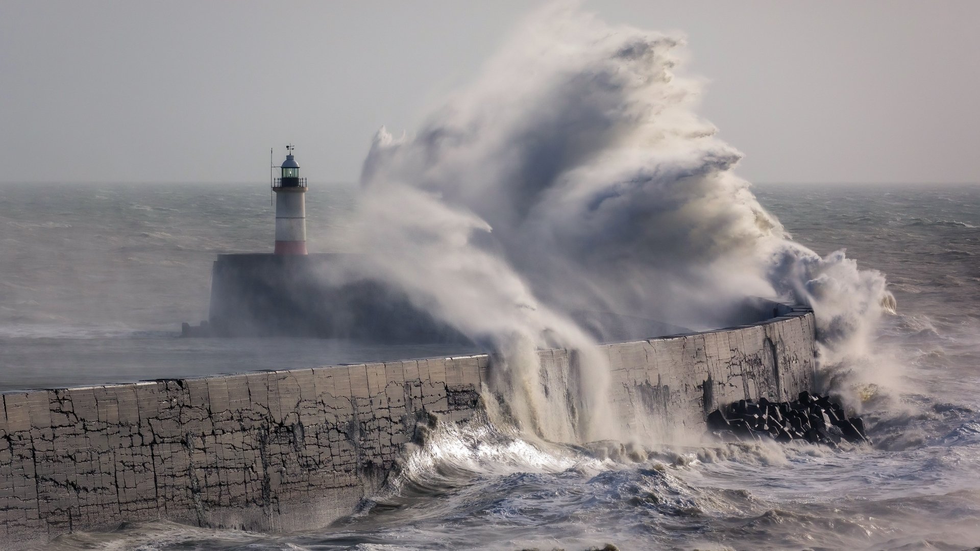 4K Ultra HD wallpaper featuring a man-made lighthouse standing strong as massive storm waves crash against the breakwater under a gray sky.