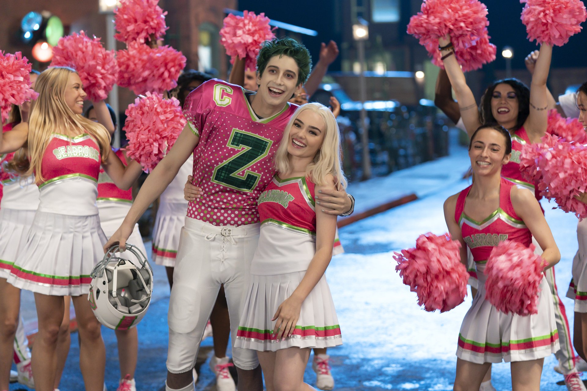 Movie Zombies 3 scene: cheerful football player in pink Z jersey embraces blonde cheerleader amid pom‑pom squad — 2K Quad HD PC desktop wallpaper/background.