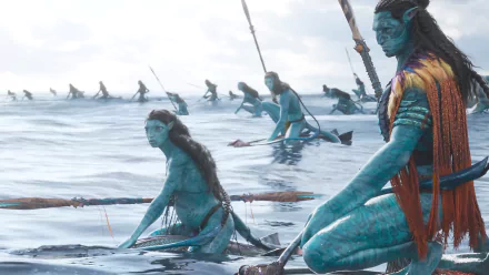 HD PC wallpaper from the movie Avatar: The Way of Water showing blue-skinned Na'vi kneeling and paddling in shallow ocean waters, with spears and boats along the horizon.