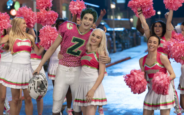 Movie Zombies 3 scene: cheerful football player in pink Z jersey embraces blonde cheerleader amid pom‑pom squad — 2K Quad HD PC desktop wallpaper/background.